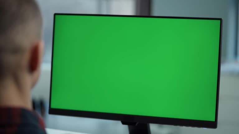 Young Man Working At Computer With Green  Mock Up Screen  Sitting At Desk in Office