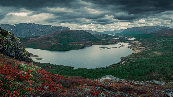 Mountain landscape with lake Nedre Leirungen from above the hike to Knutshoe summit in Jotunheimen National Park in Norway, mountains of Besseggen in background, dark cloudy sky, red moss in foreground