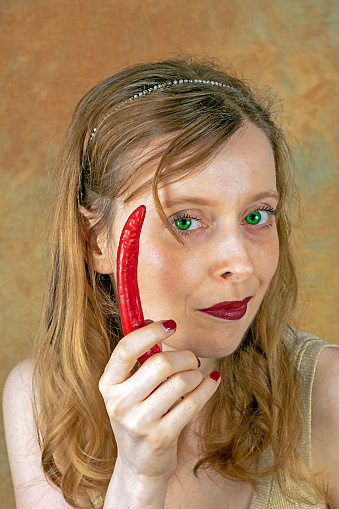 Young blonde woman with devilish neon green eyes holding spicy red chilli pepper