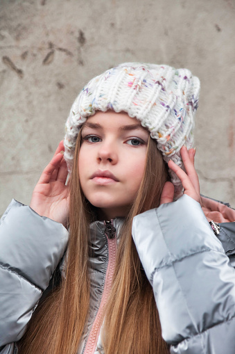 Winter portrait fashionable teenager girl in winter fashion hat posing and looking at camera at grey wall background. Positive teenage emotion outdoors. Copy text space for advertisement