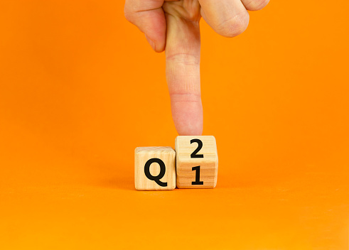 From 1st to 2nd quarter symbol. Businessman turns a wooden cube and changes words 'Q1' to 'Q2'. Beautiful orange table, orange background. Business, happy 2nd quarter Q2 concept, copy space.