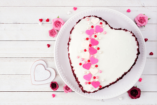Valentines Day theme red velvet heart shaped cake with white icing and colorful heart sprinkles. Above view over a white wood background.