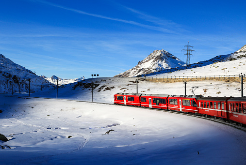 Berina, Switzerland - January 06. 2023: The red Rhaetian Rail is passing along the fronzen Lake Bianco in a dramatic sun shine hole, with the Alps summit Piz Lagalb at the background