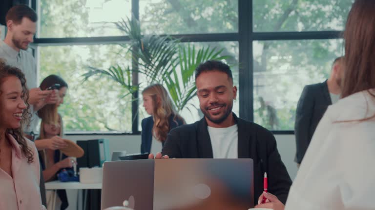 Young African American male boss enters office meeting with laptop, sits down at table to work with partners slow motion