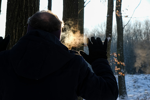 Man, with vapor coming from his mouth, in a forest on a cold sunny frosty day. Raised gloved hand.