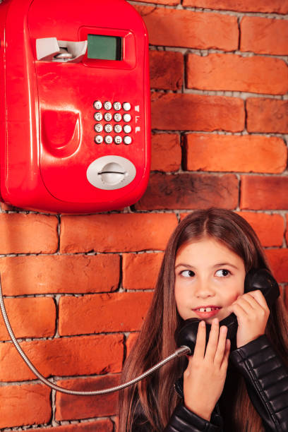 drôle de modèle de petite fille en veste en cuir appelant le vieux téléphone public rouge sur fond de mur de briques, regardant au loin. jolie enfant qui parle téléphone. concept de communication. copier l’espace de texte pour la publicité - pay phone brick wall telephone old photos et images de collection