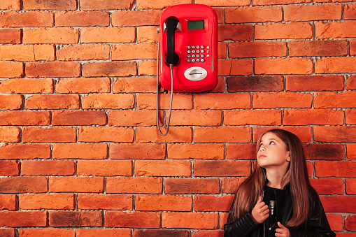 Funny little girl in leather jacket looking on old red payphone at brick wall backdrop. Small brunette lady model looking up on telephone. Communication concept. Copy space for advertising text