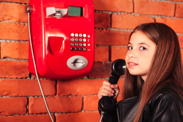 drôle de fille en veste en cuir tenant un vieux téléphone public rouge en toile de fond de mur de briques, regardant la caméra. petit modèle souriant avec téléphone. concept de communication. espace de copie pour le texte publicitaire - pay phone brick wall telephone old photos et images de collection