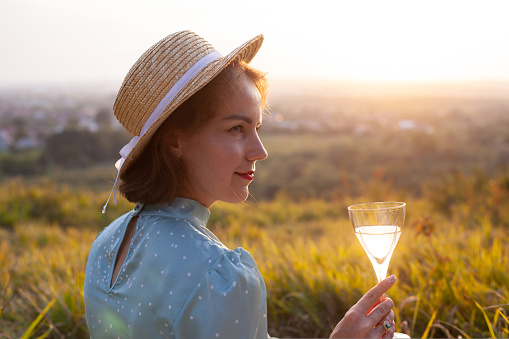 A woman in a blue dress and straw hat with short hair sitting on a white blanket holding glass and drinking white wine. Concept of having picnic in a city park during summer holidays or weekends.