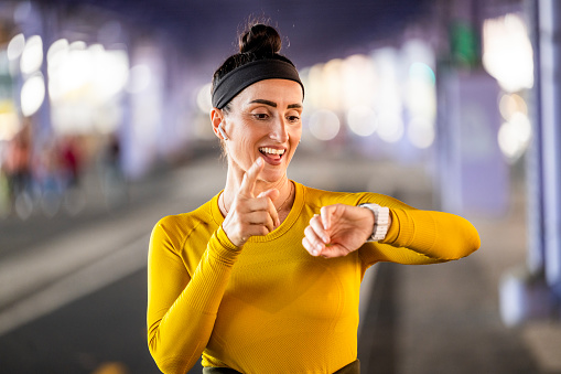 Portrait of a fit and happy woman in yellow t-shirt with a black headband and headphones while looking at her smart watch and being happy because of great fitness score during her sport activity in Manhattan.