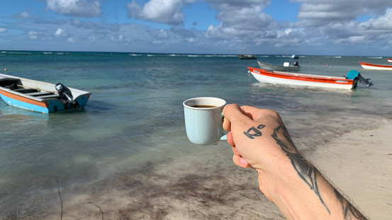 A closeup of a male hand with a tattoo holding a cup of coffee on the beach