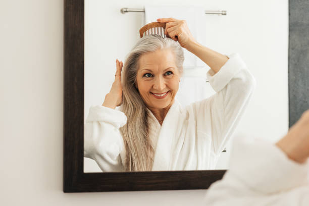 Smiling senior woman combing her grey hair in front of a bathroom mirror stock photo