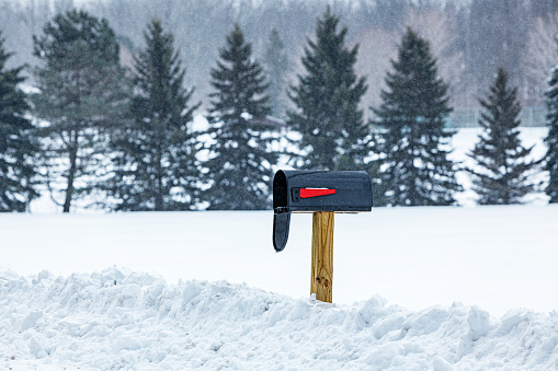 Open black painted mailbox with a bright red notification flag on a wooden post alongside a remote rural road during a winter snow storm.