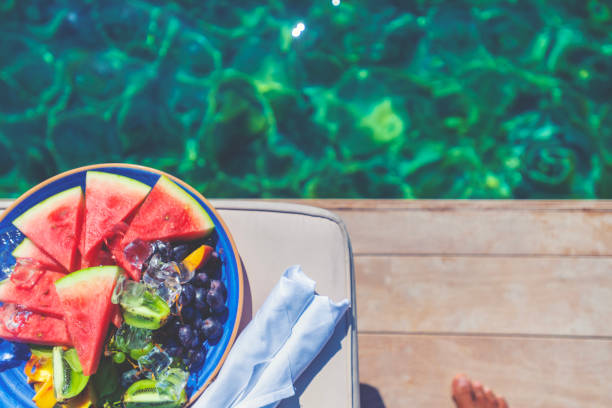 Plate of watermelon and fruit on a deck by the ocean. The water is crystal clear turquoise. Sunny clear day. A mans foot can be seen