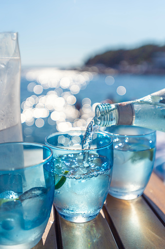 Pouring water from a bottle into a glass at the beach. Lemon and ice in the blue glasses. Backlit at sunset