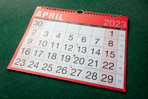 Calendar 2023, April, monthly planner. Day, month, year, date and activity organiser wall and desk planner. Red and white calendar with large letters and numbers on green background. Shown at angle.