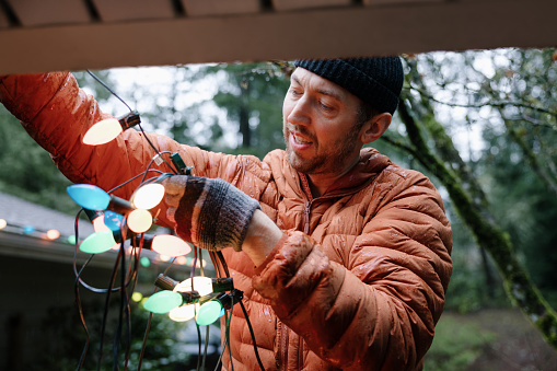 A middle aged Caucasian man works on his Christmas holiday decor, either putting up or taking down the colored light bulb decorations from off of his house gutters.  Shot on a rainy day in Washington state.