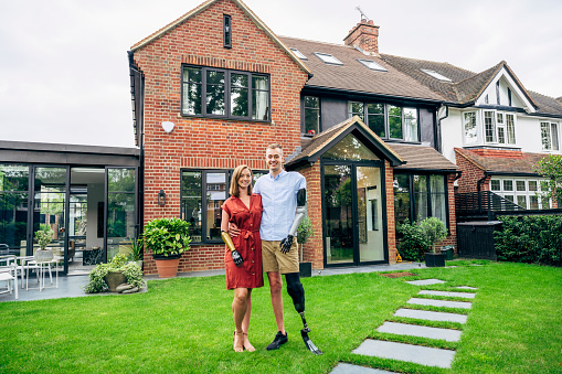 Full length view of Caucasian couple wearing prosthetics and standing together in back yard of modern, semi-detached brick house smiling at camera.