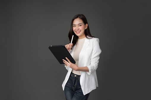 Female executive wearing a white coat with long sleeves Taking photos against gray background in the studio. She holds a laptop tablet and a pen. Stand up and make eye contact and smile beautifully