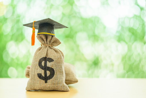 Education or student loan, financial aid and scholarship concept : Mortarboard or graduation cap on top of a US dollar cash bag. Depicting financial assistance for a student's tuition and other costs.