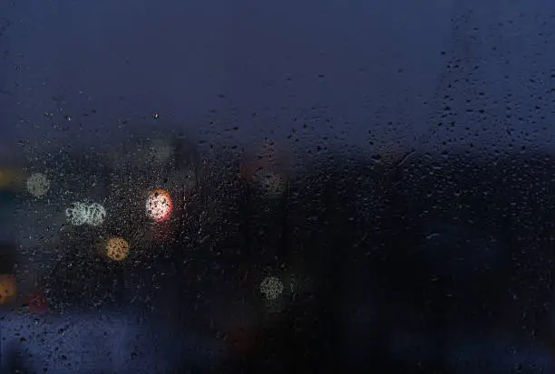 Photo of Raindrops on a window glass and unfocused lights of a evening city. Background of night city behind glass during rain, copy space