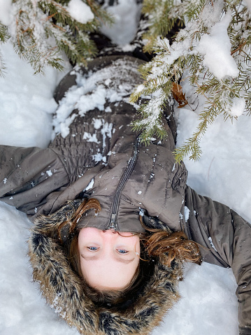Beautiful teen girl, laying down and playing in the snow under a pine tree. She is laying upside down and looking at the camera, wearing a furry hooded winter coat and snow pants. Taken in Northern Wisconsin, USA.