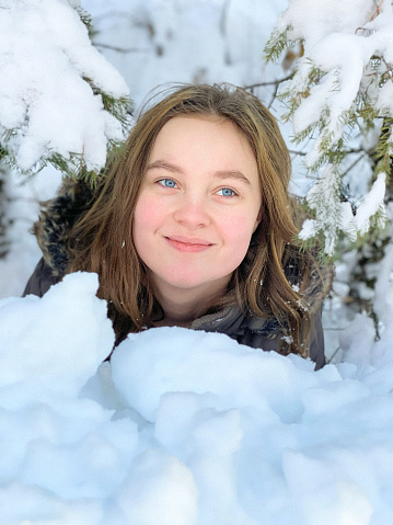 Beautiful teen girl, smiling and laying down and playing in the snow under a pine tree. She is laying down on her stomach, her head up, looking away from the camera and looking like she’s having fun and playing. Taken in Northern Wisconsin, USA.
