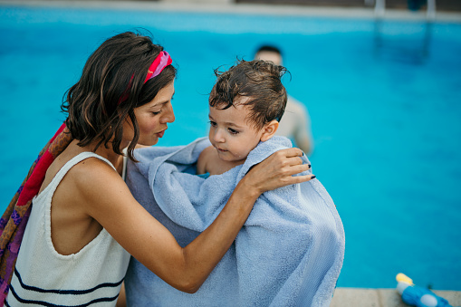 Dedicated and caring mother wiping her wet toddler after pool time. Father in the background.