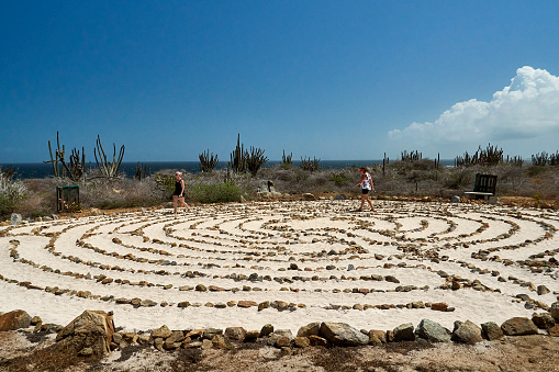 Noord, Venezuela – February 22, 2020: Two ladies walking the sandy Aruba Peace Labyrinth at the Alto Visto Church with a view of the ocean, cactus and a bright blue sky.