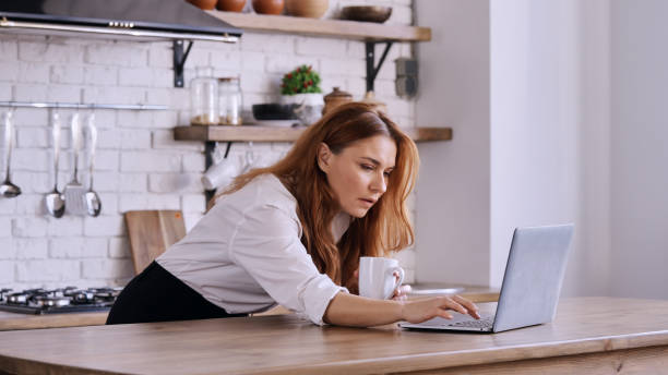 istock Caucasian female working on her laptop in a kitchen and drinking coffee 1454964444