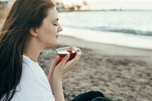 Young woman having picnic, drinking hot fresh tea sitting on the beach at sunset.