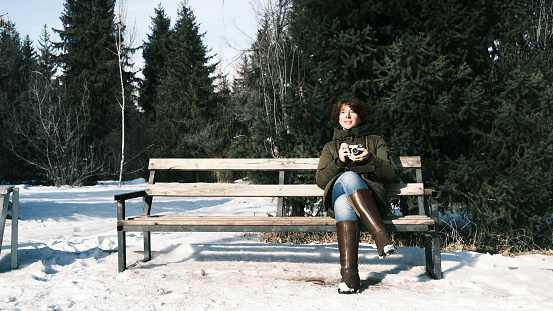 Woman is using her vintage film camera while sitting on sunny winter park bench