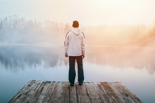 Young man in hoodie, hat and pants standing on wooden pier on pond shore with melancholy fog at sunrise. Czech morning landscape