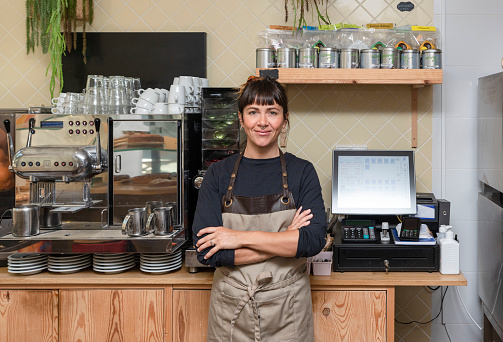female business owner behind the counter of a coffee shop with crossed arms, looking at camera.
