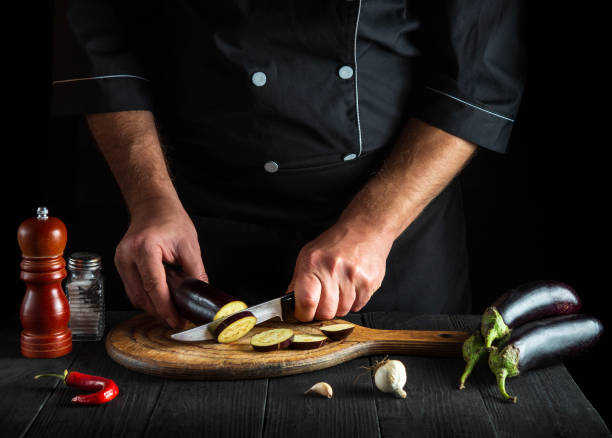 professioneller koch schneidet eine aubergine in der restaurantküche. nahaufnahme der hände des kochs während der arbeit. leckeres frühstück oder abendessen - eggplant group of objects raw food eating stock-fotos und bilder