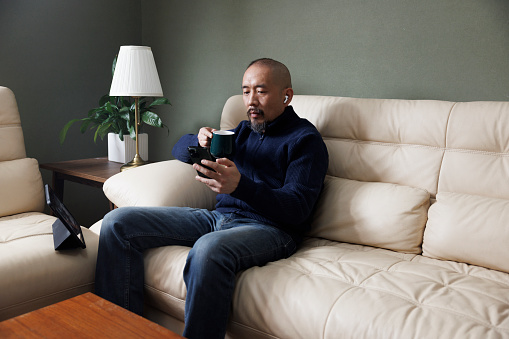 This is a middle-aged Asian man, about 40 years old. Wearing a blue sweater, sitting in the living room at home. While drinking coffee, he used his mobile phone to read information.