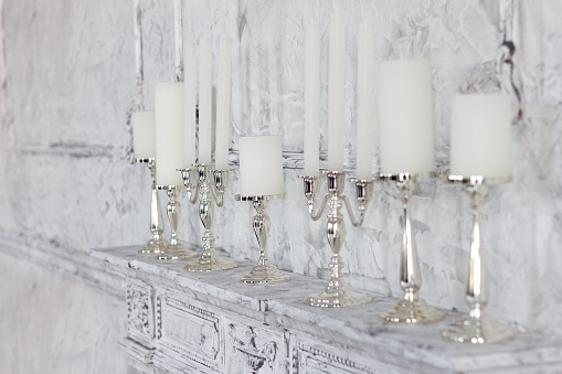 Elegant interior, white candles in elegant candlesticks stand on a fireplace in the room. Focus on fourth candlestick