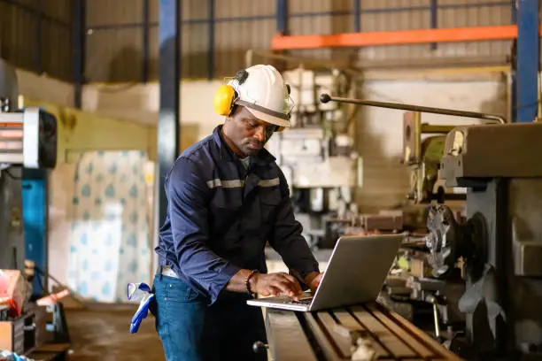 Photo of Male technician specialist with safety uniform using laptop checking and controlling machinery at manufacturing plant