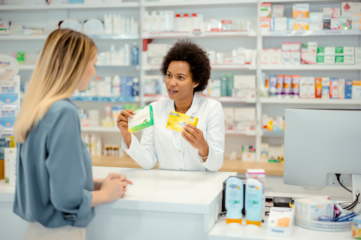 Pharmacy: Professional African American Pharmacist Helping Young Female Customer with Medicine Recommendation, Advice, Talking. Drugstore with Full of Drugs, Pills, Health Care