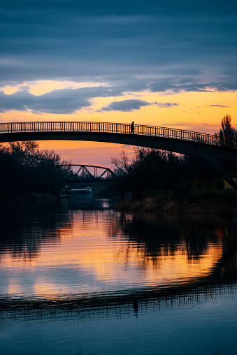 Silhouette of a man walking across small bridge over a peaceful river at sunset. Serene scenery in Zrenjanin city in Serbia on the river Begej