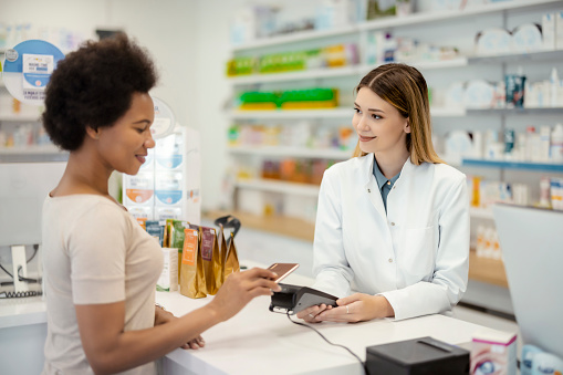 Pharmacy Drugstore Checkout Cashier Counter: Pharmacist and a Customer Using NFC Smartphone with Contactless Payment Terminal to Buy Prescription Medicine, Health Care Goods