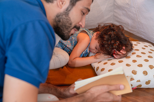 happy father is reading a fairytale book to toddler daughter child girl sleeping lying on the floor in living room at home. bedtime story Children sleeping near  father with book on soft pillow