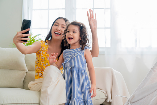 happiness lifestyle at home ,asian female woman nanny aupair takecare playing smartphone selfie with little child girl with cheerful laugh smiling in living area at home morning weekend peaceful