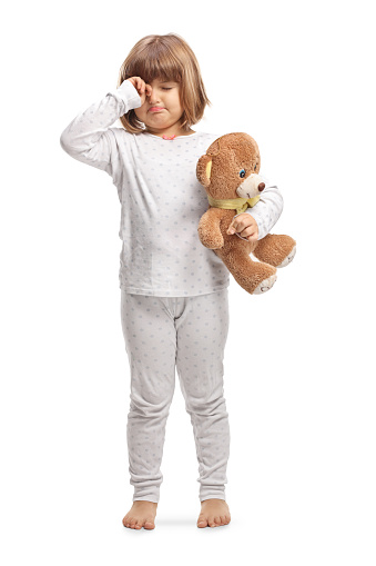 Child in pajamas holding a teddy bear and rubbing eyes isolated on white background