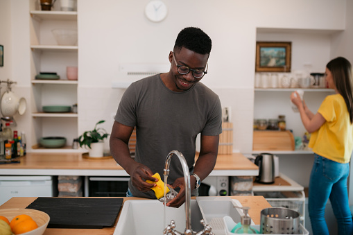 Young african man washing dishes in sink with female friend working in background in kitchen. Multiracial roommates doing chores in kitchen at home.