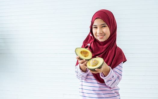 Little sweet girl wearing hijab or headscarf smiling with happiness, posing, holding avocado for health with copy space. Religion, Education Concept