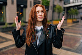 Portrait of frustrated red-haired young woman touching wet hair after autumn rain standing on beautiful city street.