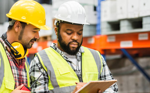 istock Two diverse professional male workers holding board, checking shipping stocks in storage, warehouse or factory for delivery, wearing safety hat. Diversity, Commercial Industry Business Concept. 1455040193