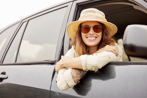 Portrait of a woman hanging out the window of a car while taking a road trip, travelling to her destination. Attractive young female hipster smiling and looking relaxed, wearing a hat and sunglasses