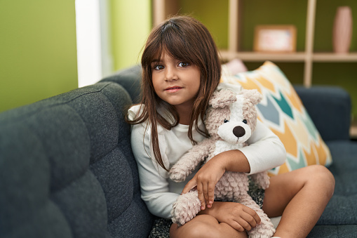 Adorable hispanic girl hugging teddy bear sitting on sofa at home